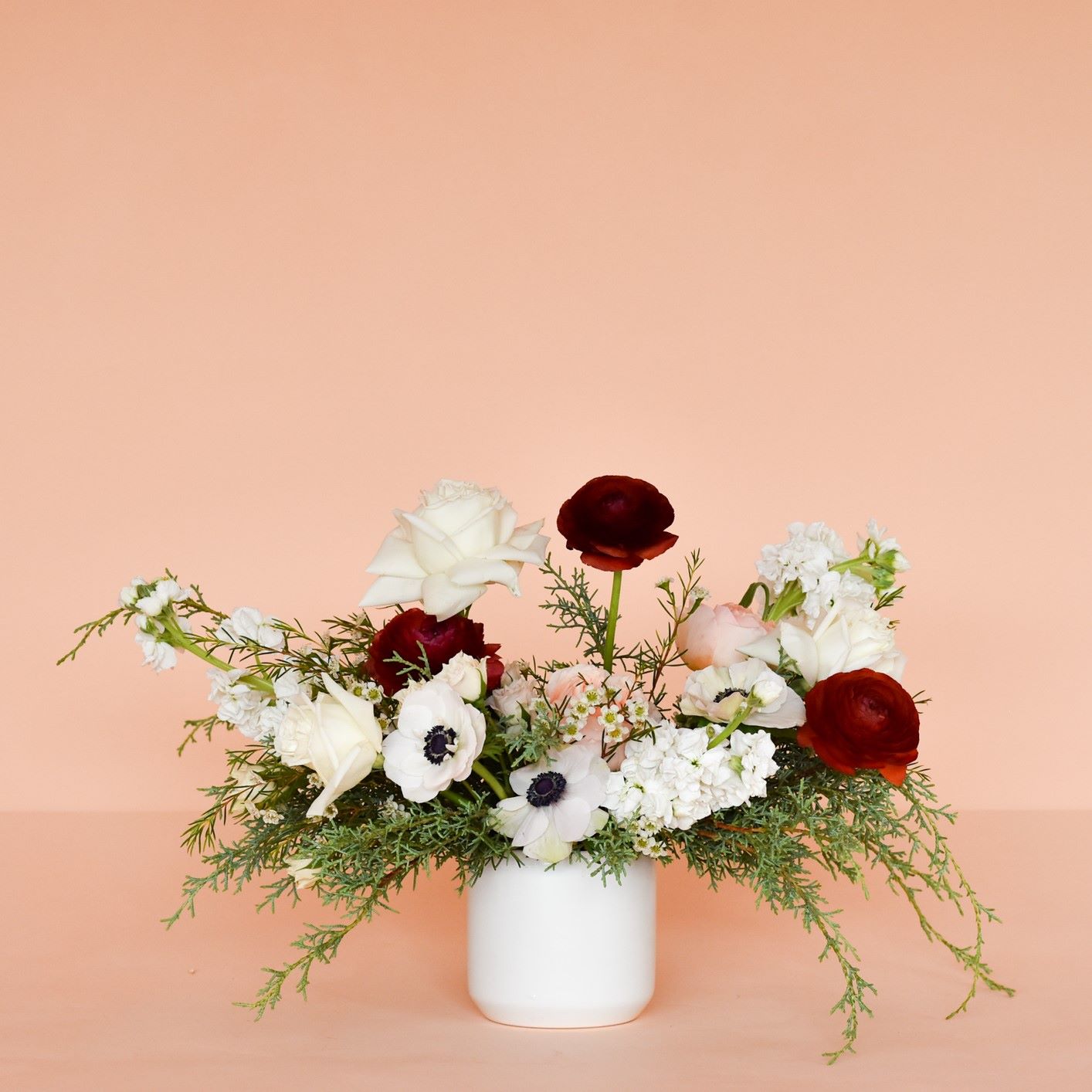 A holiday centerpiece in a low, simple white vase with ranunculus, stock, roses, cosmos, winter greens and more