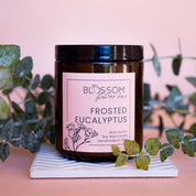 An amber jar candle with Blossom's label for Frosted Eucalyptus with eucalyptus in the background