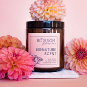 An amber glass candle with Blossom's label for Signature Scent. The candle is surrounded by peach colored dahlias. 